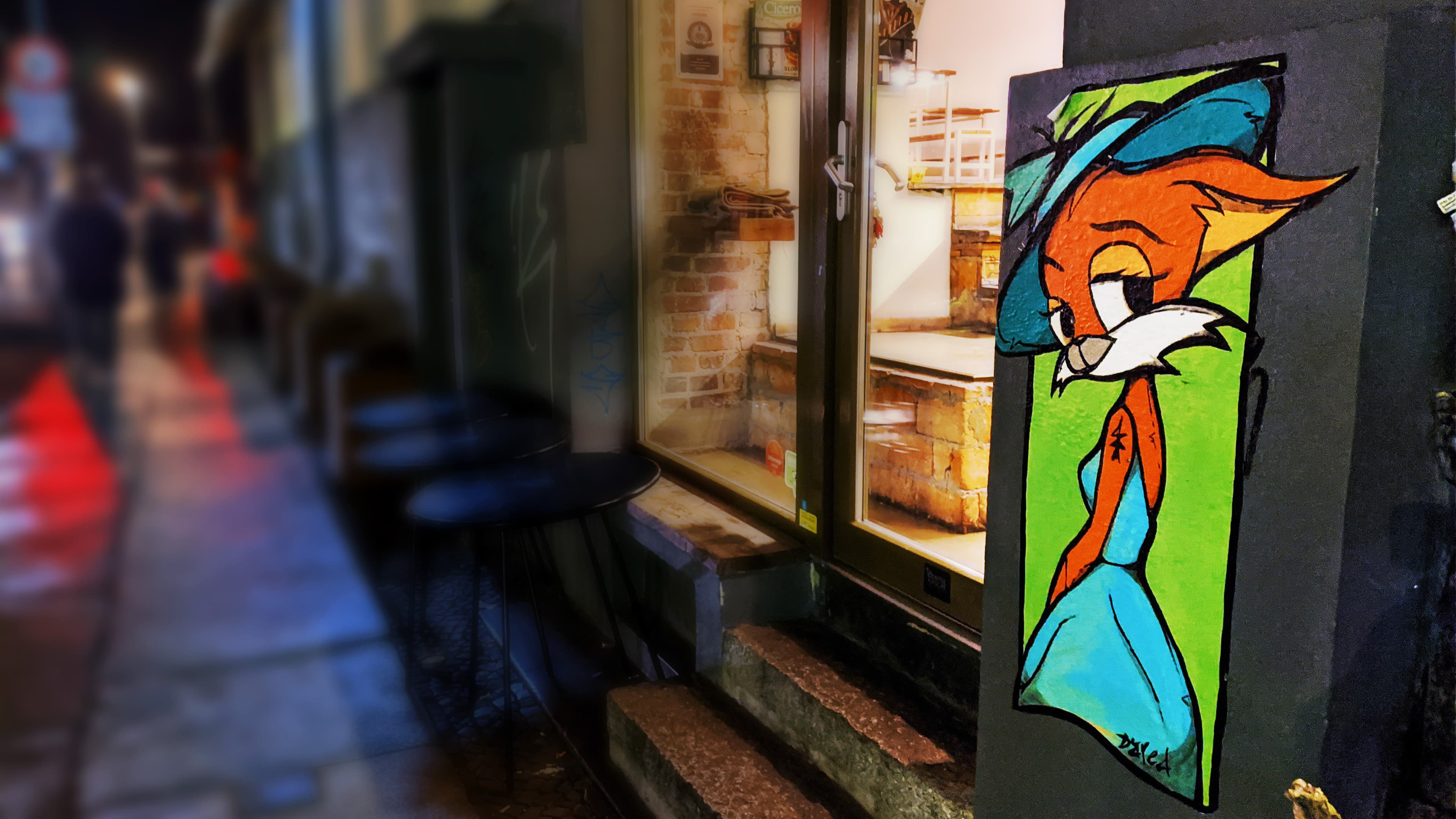 Night scene in Berlin featuring a detailed paste-up of 'Faye the Fox' by street artist Dared. The anthropomorphic fox wears a blue dress and hat against a green backdrop, placed on a pillar next to a glowing shop window and blurred cafe seating.