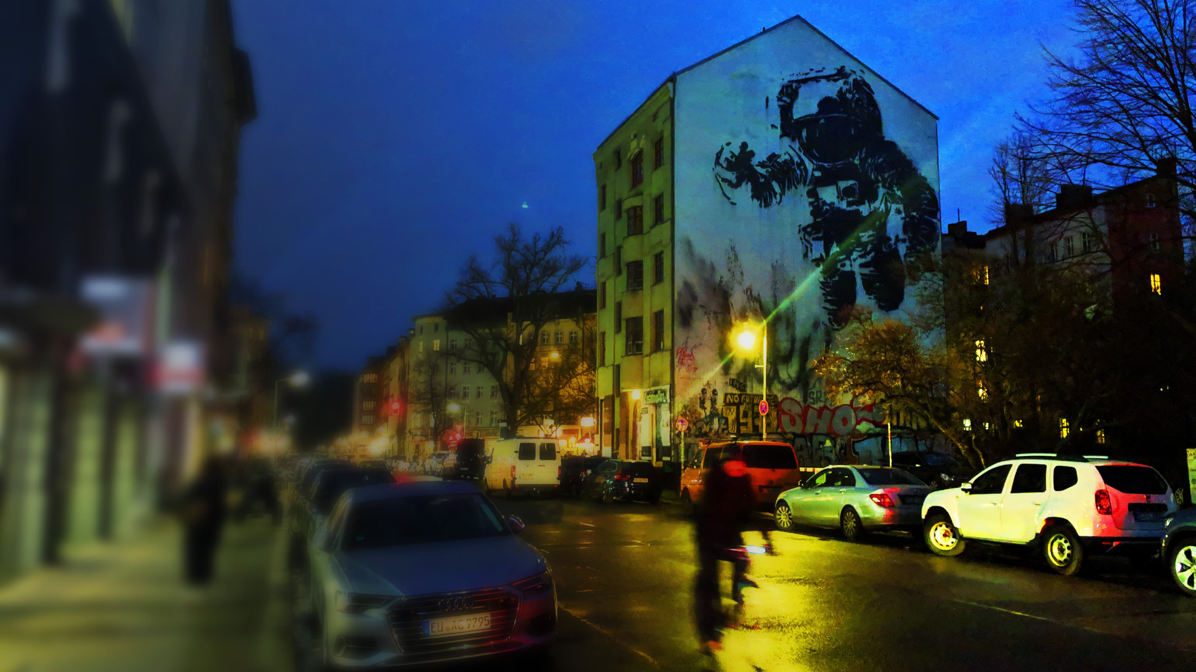 Night view of a residential building in Berlin-Kreuzberg featuring the iconic large-scale mural 'Astronaut Cosmonaut' by Victor Ash. The stencil-style artwork depicts a floating astronaut in black and white, towering over the urban cityscape.