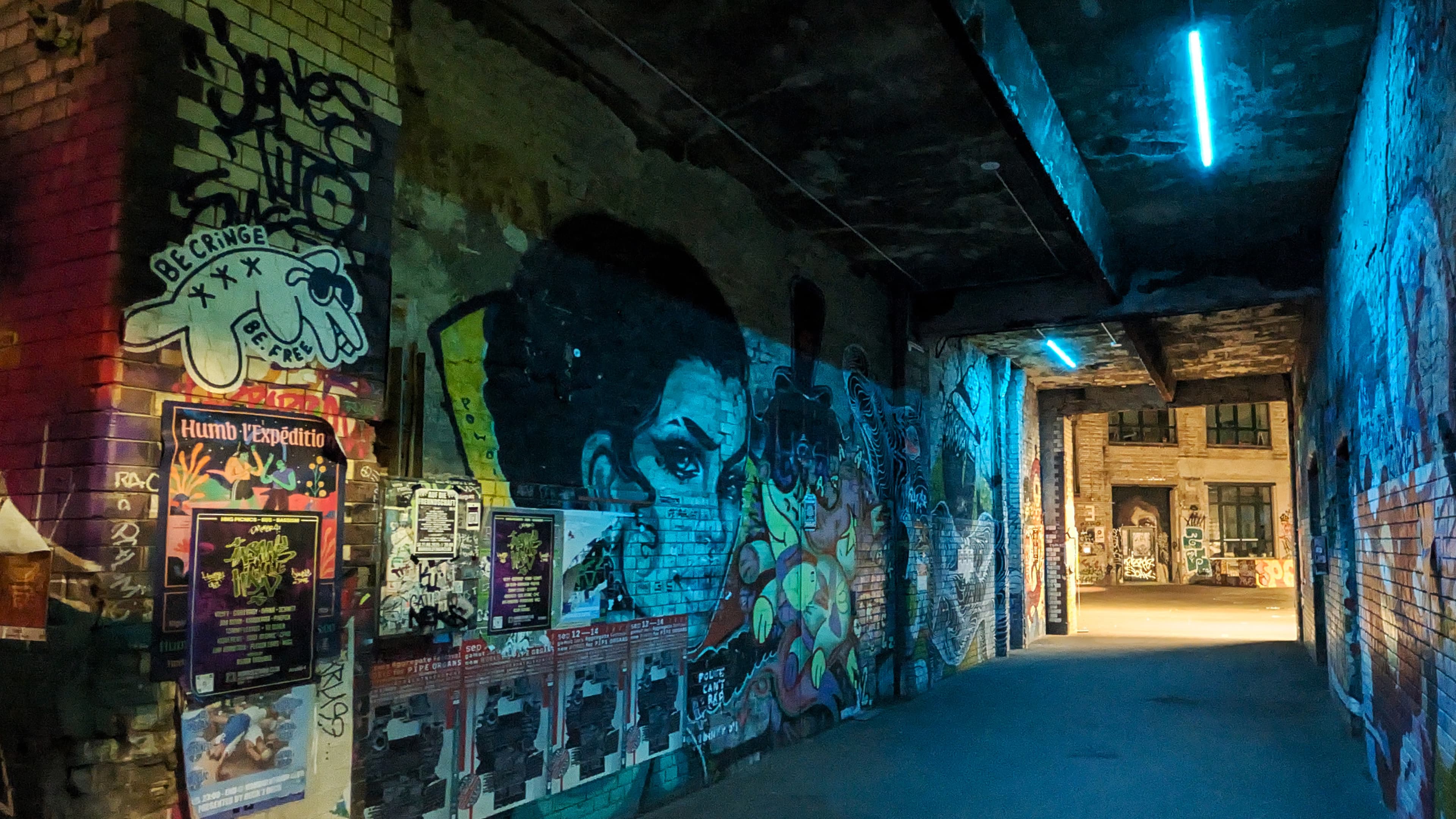 A vibrant brick wall in a Berlin courtyard at night, heavily covered with various graffiti tags, stickers, and urban posters. The scene captures the raw subculture of the city, showcasing layered street art characters and colorful textures.