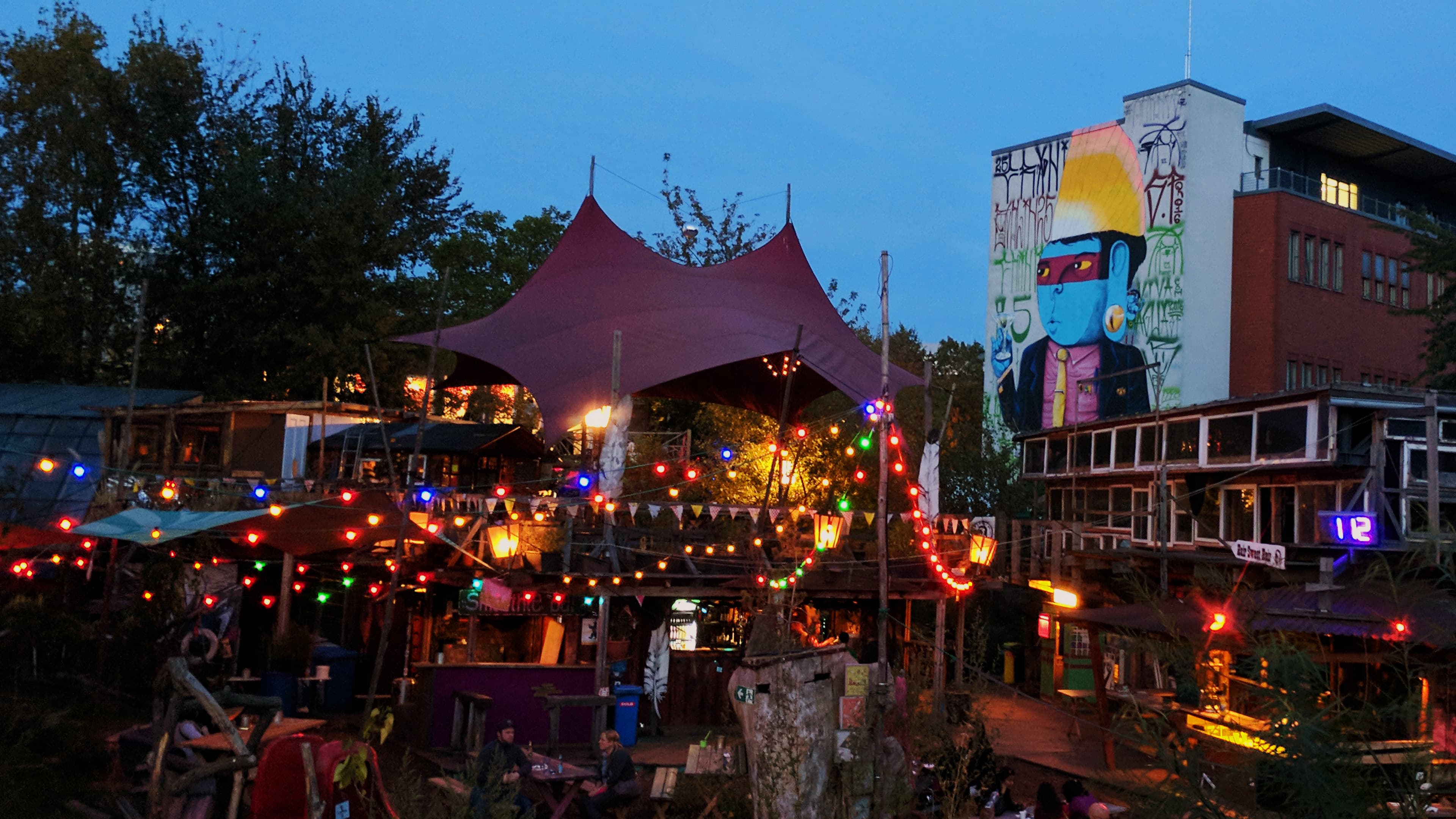Atmospheric evening at Holzmarkt 25 by the Spree in Berlin, showing the 'Pampa' area with fairy lights and a sun sail. In the background, the colorful mural 'Big Business!' by artist Cranio is visible on a creative residential and studio building.
